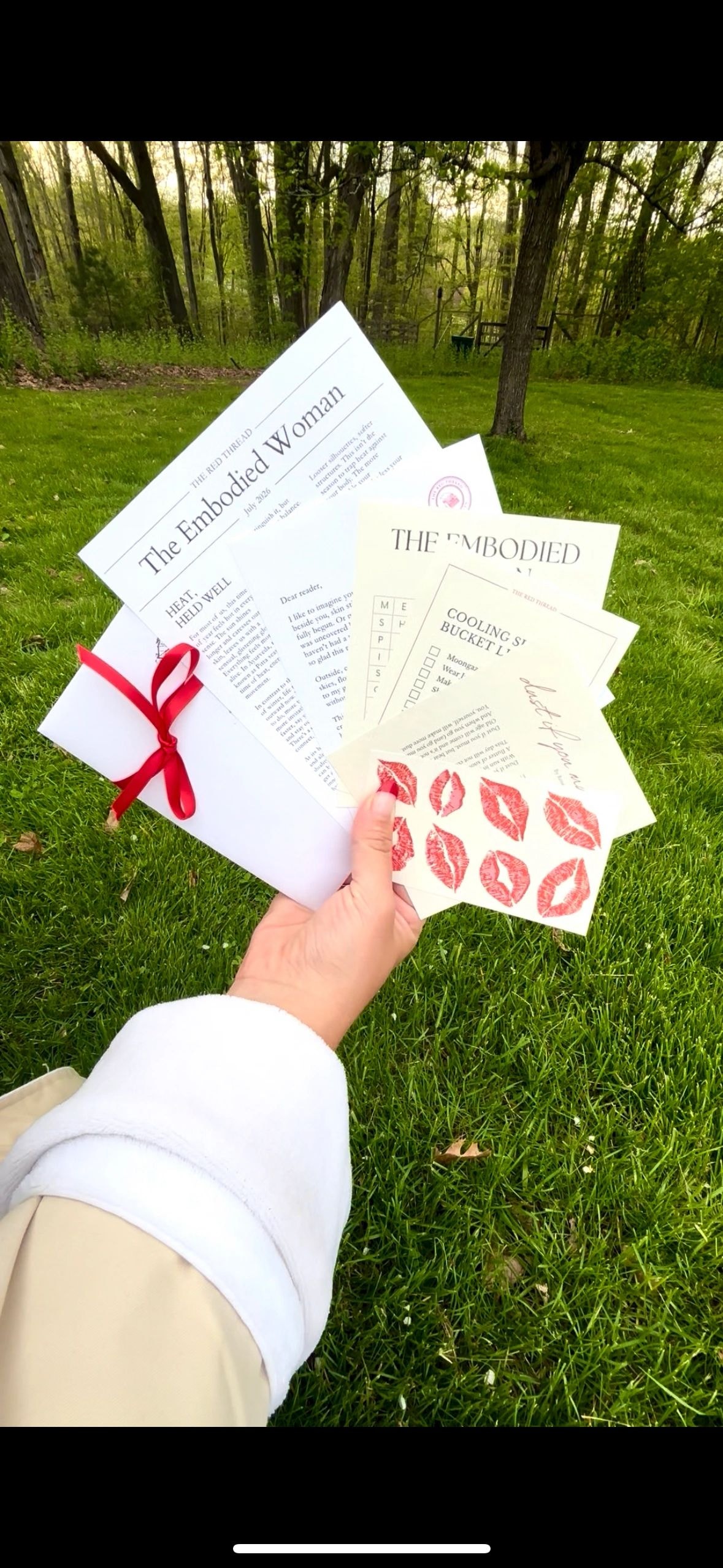 Hand holding fanned-out Embodied Woman letters tied with red ribbon against green grass